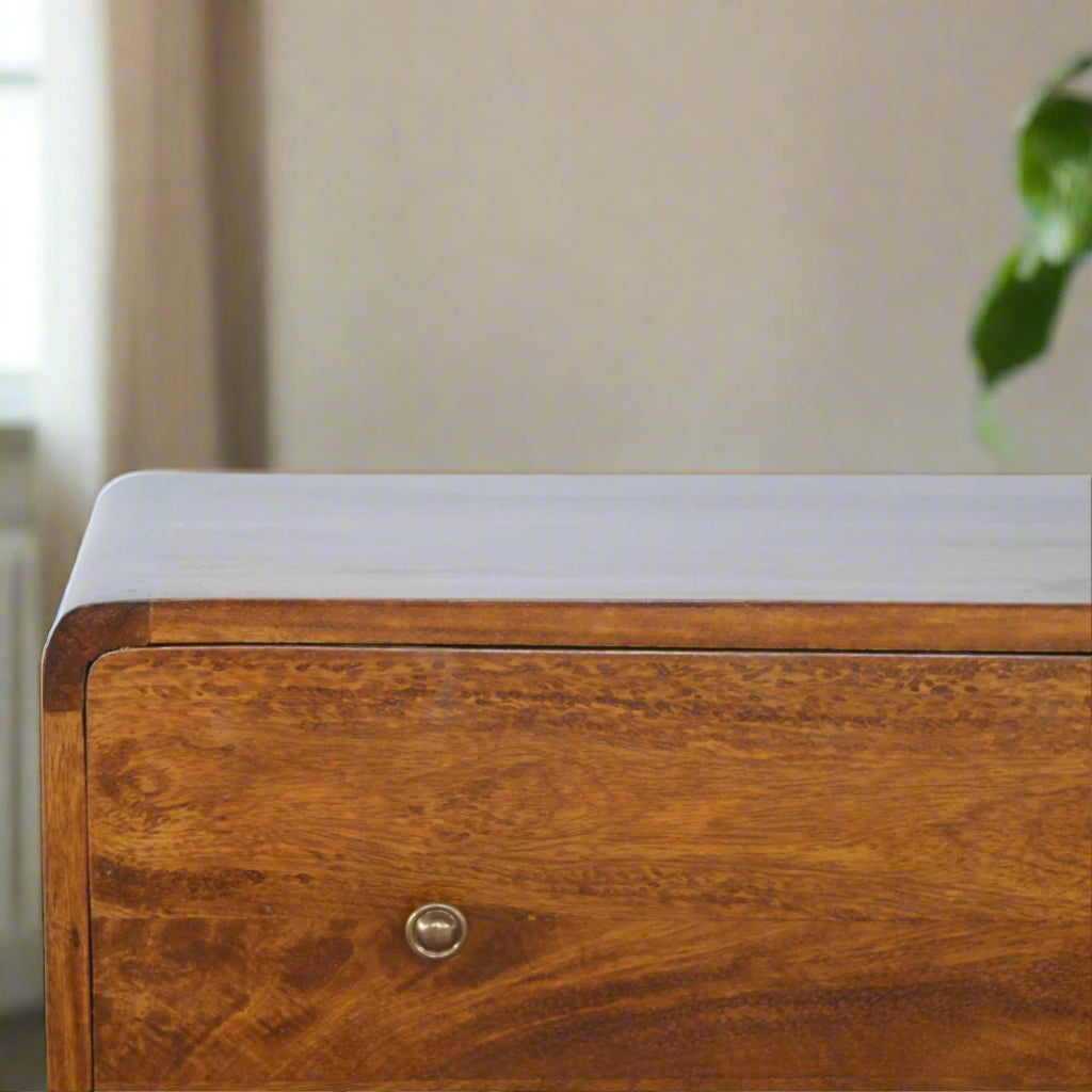 "Curved chest of drawers made from solid mango wood with chestnut finish, featuring shiny accent knobs, four Nordic-style legs, and smooth, rounded corners for a modern, contemporary look."